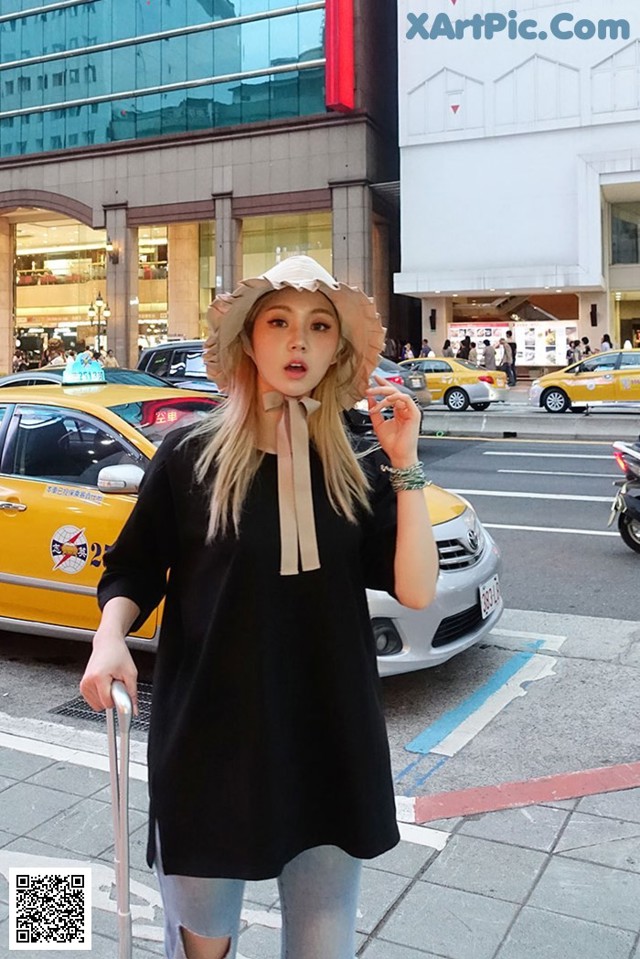 A woman standing on the sidewalk with a suitcase and a hat.