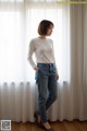 A woman standing in front of a window wearing a white shirt and jeans.